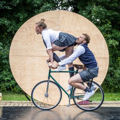 Twee mensen op een fiets. De voorste persoon staat voorovergebogen op het frame, terwijl de achterste persoon op de bagagedrager zit.