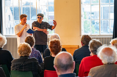 Twee mannen presenteren op het podium voor een publiek in een lichte zaal met grote ramen.