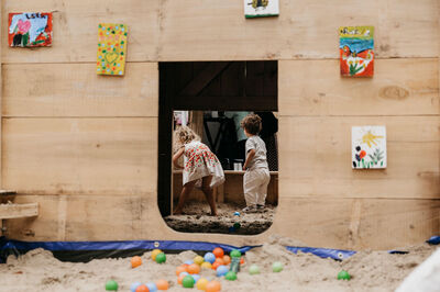 Kinderen spelen in een houten speelhuisje met kleurrijke schilderijen op de muren en een zandbak eromheen.