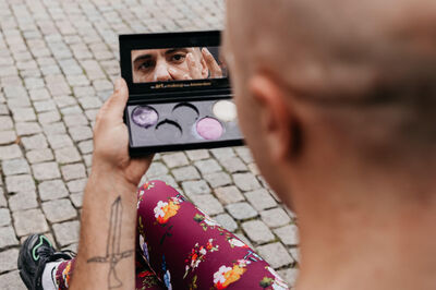 Man met een kaal hoofd kijkt in een spiegel van een make-uppalette, met kleurrijke tinten zichtbaar op de lippenstift.
