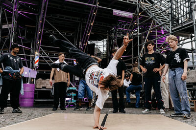 Een breakdancer voert een handstand uit op een podium, omringd door een enthousiaste menigte.