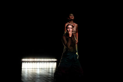 Twee danseressen op een donkere achtergrond, één staat achter de ander met een dramatische pose. Verlichting uit het midden.