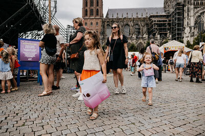 Kinderen lopen met een groot werkblad over een plein. Volwassenen lopen ook op het plein.