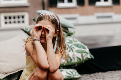 Een meisje met een bloemenhoofdband kijkt door haar handen als een verrekijker, zittend op een zachte ondergrond.