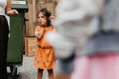 Een meisje in een oranje jurk staat op de straat, met een groen afvalcontainer op de achtergrond.