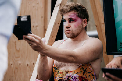 Man in een kleurrijke jurk kijkt in een spiegel, met opvallende make-up en een zelfverzekerde uitdrukking.