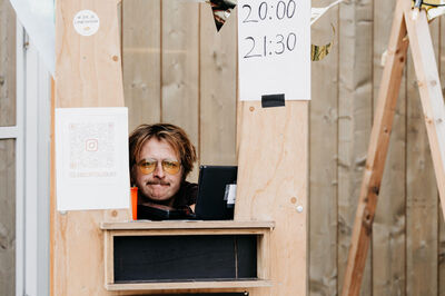 Een persoon zit achter een houten balie met een laptop, omringd door een houten omheining en aankondigingen.