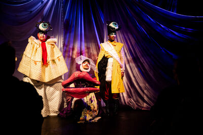 Drag performers in elaborate costumes pose on stage against a colorful backdrop.