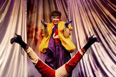 Een clown in kleurrijke kostuums staat op het podium, terwijl een andere performer ondersteboven ligt.