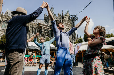 Groep mensen, waaronder twee dansers, voert een optreden uit op een plein met historische gebouwen op de achtergrond.