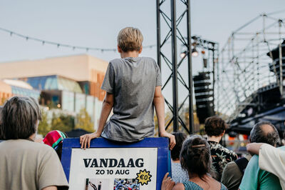 Een jongen zit op de schouders van een volwassene en kijkt naar een podium met optredens op een evenement.