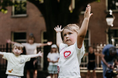Kinderen spelen buiten; een meisje met een T-shirt steekt haar armen omhoog, terwijl andere kinderen op de achtergrond zijn.