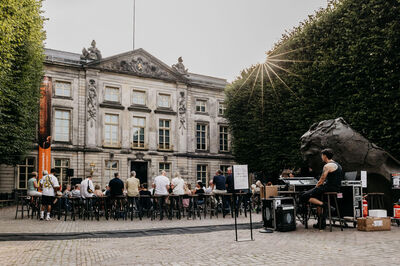 Een buiten locatie met een groep mensen die aan een lange tafel zitten, terwijl een muzikant speelt voor een historisch gebouw.