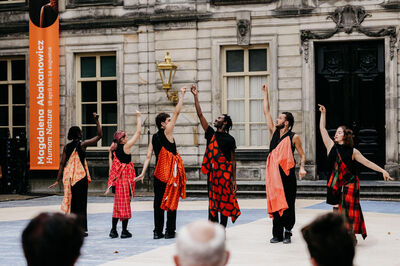 Vijf vrouwen in kleurrijke kostuums voeren een dansvoorstelling uit buiten, met een publiek op de achtergrond.