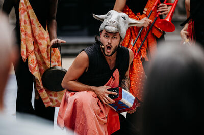Man in schapenkop en kleurrijke kleding, zittend tussen een groep muzikanten tijdens een theatrale voorstelling.