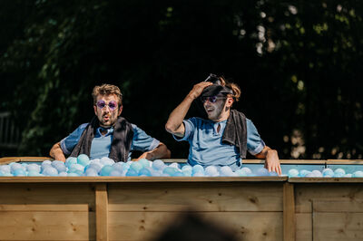 Twee mannen zitten in een houten bad gevuld met blauwe ballen, zichtbaar ontspannen met handdoeken om hun nek.