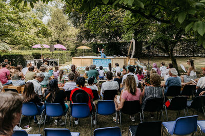 Kindervoorstelling in park met publiek dat naar een performace kijkt, omringd door bomen en kleurrijke parasols.