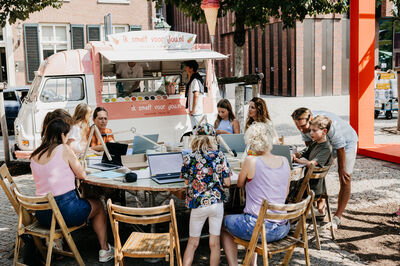 Een groep mensen zit aan een tafel buiten, met laptops, terwijl ze genieten van snacks van een mobiele foodtruck.
