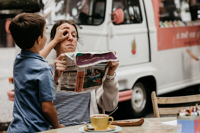 Een kind houdt de krant van een vrouw omhoog, terwijl ze met een verraste blik naar hem kijkt, met een foodtruck op de achtergrond.