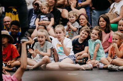 Een groep kinderen zit op de grond en kijkt enthousiast naar een voorstelling, omringd door volwassen toeschouwers.