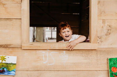 Een blije jongen kijkt uit het raam van een houten speelhuisje, met kindertekeningen aan de muur.