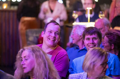 Twee personen zitten naast elkaar tussen publiek en lachen.