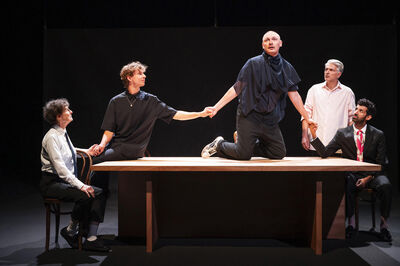 Vijf acteurs presenteren een theatrale scène rond een tafel, met één acteur staand in het midden.