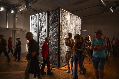 Bezoekers verkennen een kunstinstallatie met grote panels in een galerie, omringd door houten vloeren en dim licht.