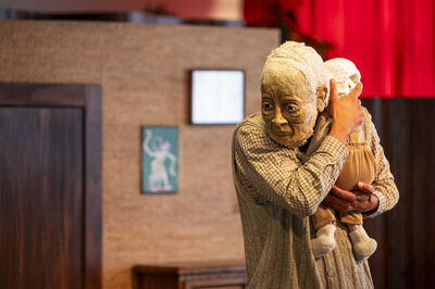 Beeld van een menselijk figuur met een kinderpop, waarbij een oudere man de pop tegen zich houdt in een interieur.