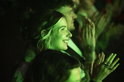 Een vrouw met een glimlach, omringd door andere mensen, applaudisseert in een donkere ruimte met groene verlichting.