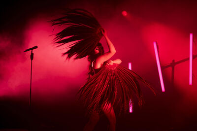 Een performer in een extravagante outfit met veren danst op een podium, omringd door rode lichten en rook.