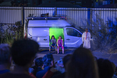 Twee kinderen zitten in een wagen met groene verlichting, terwijl een volwassene hen observeert vanuit de schaduw.