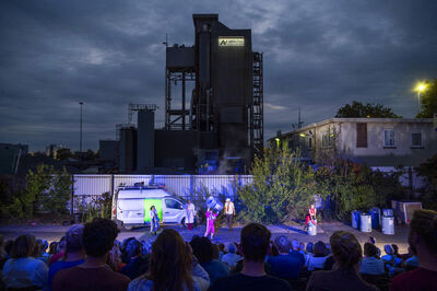 Theateroptreden in een buitenruimte, met acteurs op een podium en een industrieel gebouw op de achtergrond.