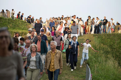 Een grote groep mensen wandelt een groene heuvel af, met een zonnige lucht op de achtergrond.