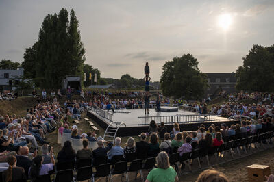 Een groot publiek kijkt naar een circusvoorstelling op een buitenterrein, met een artiest op een podium tijdens zonsondergang.