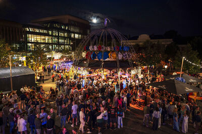 Een levendig evenement ’s nachts met een grote menigte, kleurrijke verlichting en een fontein aan het centrum.