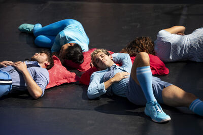 Groep dansers ligt op de vloer in verschillende houdingen, gekleed in kleurrijke kleding.