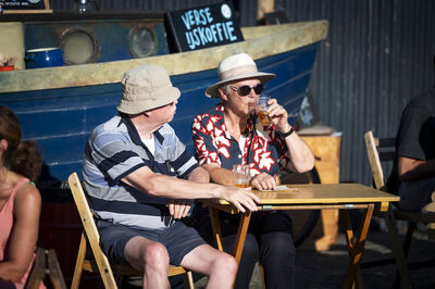 Twee mannen in zonnige kleding zitten aan een tafel bij een houten boot en drinken iets.