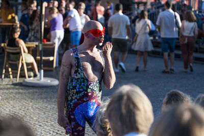 Een performer in kleurrijke kleding stelt zich voor een publiek voor op een drukke buitenlocatie.