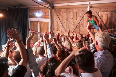 Een energieke live muziekoptreden met een artiest op het podium en enthousiaste bezoekers die dansen en hun handen in de lucht steken.