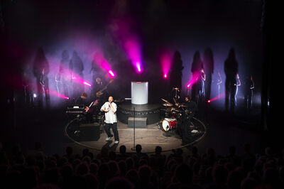 Een muzikant performt op een podium, omgeven door kleurrijke lichten en schaduwen van andere muzikanten.