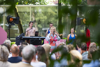 Zangeres treedt op voor een publiek met muzikanten op een buitenpodium, omringd door groene bladeren.