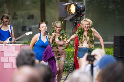 Drie vrouwen in kleurrijke kleding en met bloemen in het haar dansen op een podium, terwijl een muzikant gitaar speelt.