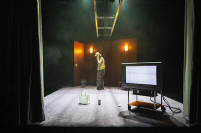 Een persoon staat in een verlaten theater met een tv en deuren, omgeven door lichteffecten en rook.