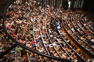 Een volle zaal met mensen die in een cirkelvormige opstelling zitten, uitzicht van bovenaf.