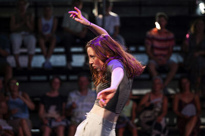 Een danseres voert een expressieve beweging uit op een podium, met een publiek op de achtergrond.