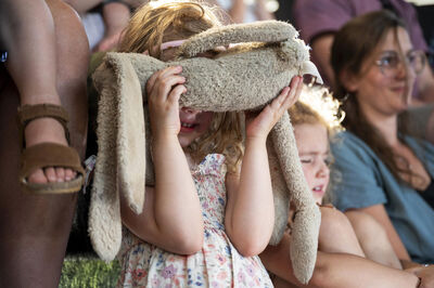 Meisje met een knuffel dat haar gezicht bedekt, terwijl ze naar een voorstelling kijkt, omringd door andere toeschouwers.