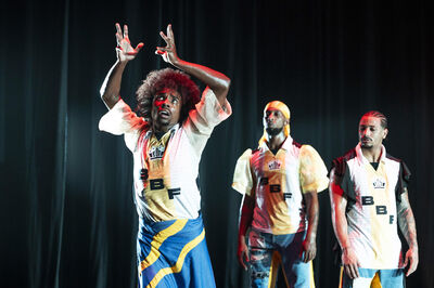 Groep dansers in kleurrijke kostuums voert een energievolle dansroutine op een podium met theatrale verlichting.