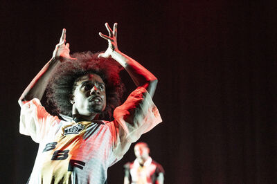 Een danser met afro haar voert een krachtige dansroutine uit op een podium met een rode achtergrond.