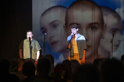 Twee muzikanten op een podium met een projector die gezichten toont in de achtergrond. Mensen zijn in de schaduw zichtbaar.